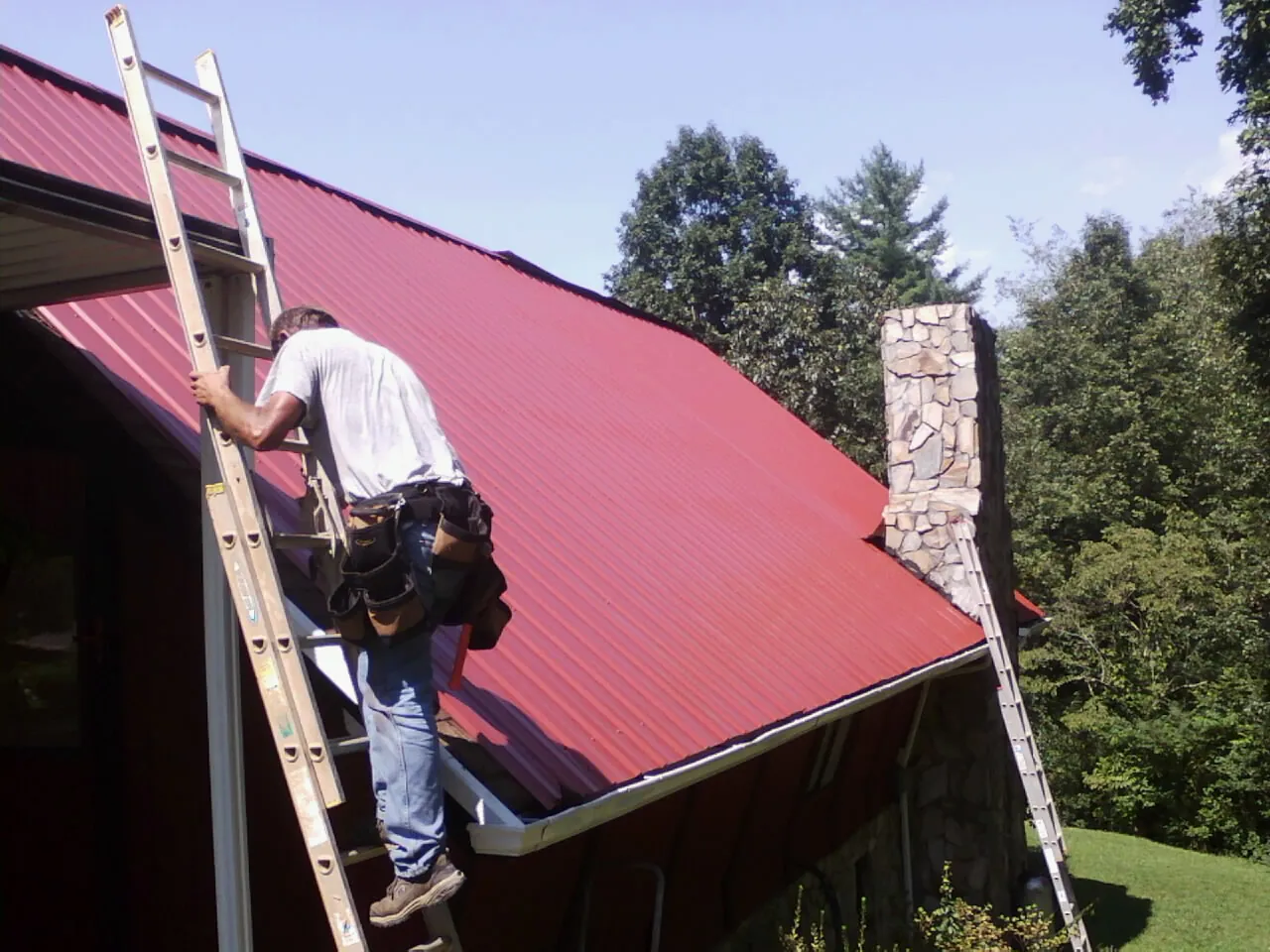 Roofer on a ladder beside a red metal roof and stone chimney — Soffit & Fascia Repair by Roofing Brothers in Waldorf